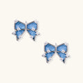 Blue denim bow stud earrings with white gold plating and crystal accents on cream background