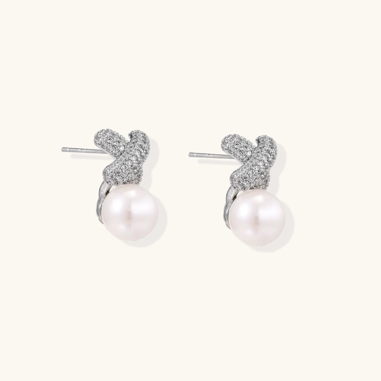 White gold plated studs featuring pavé X-shaped tops with freshwater pearl drops on cream background.